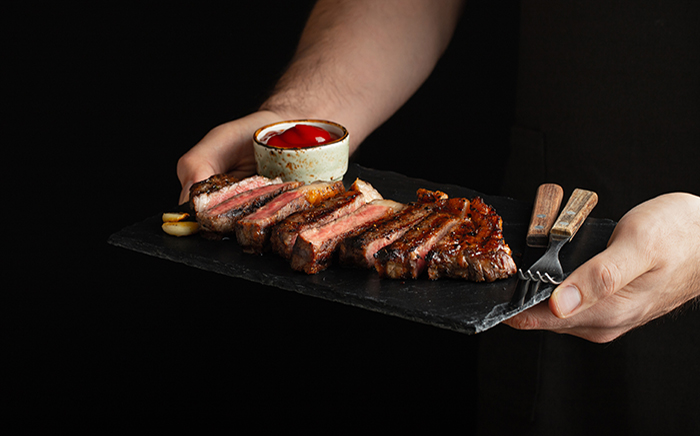 Man holding a slate plate with a sliced beef steak, sauce, a knife and a fork