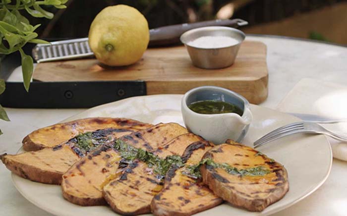 Boniatos en un plato con aliño de cilantro y aceite con una tabla de madera con un limón detrás en una barbacoa vegana