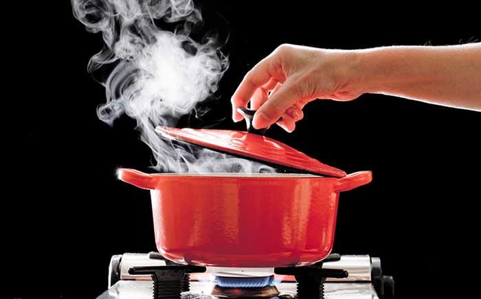 Cacerola de color rojo puesta al fuego cocinando mientras una mano levanta la tapa